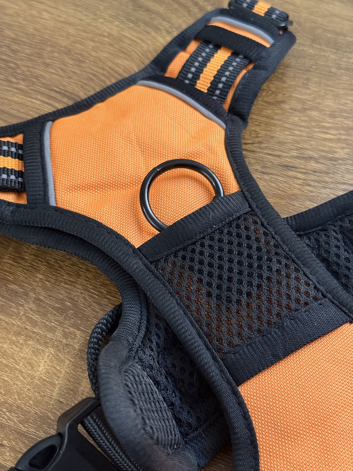 Close-up of an orange and black dog harness laid flat on a wooden surface, showing mesh panels, adjustable straps, and a black metal ring.