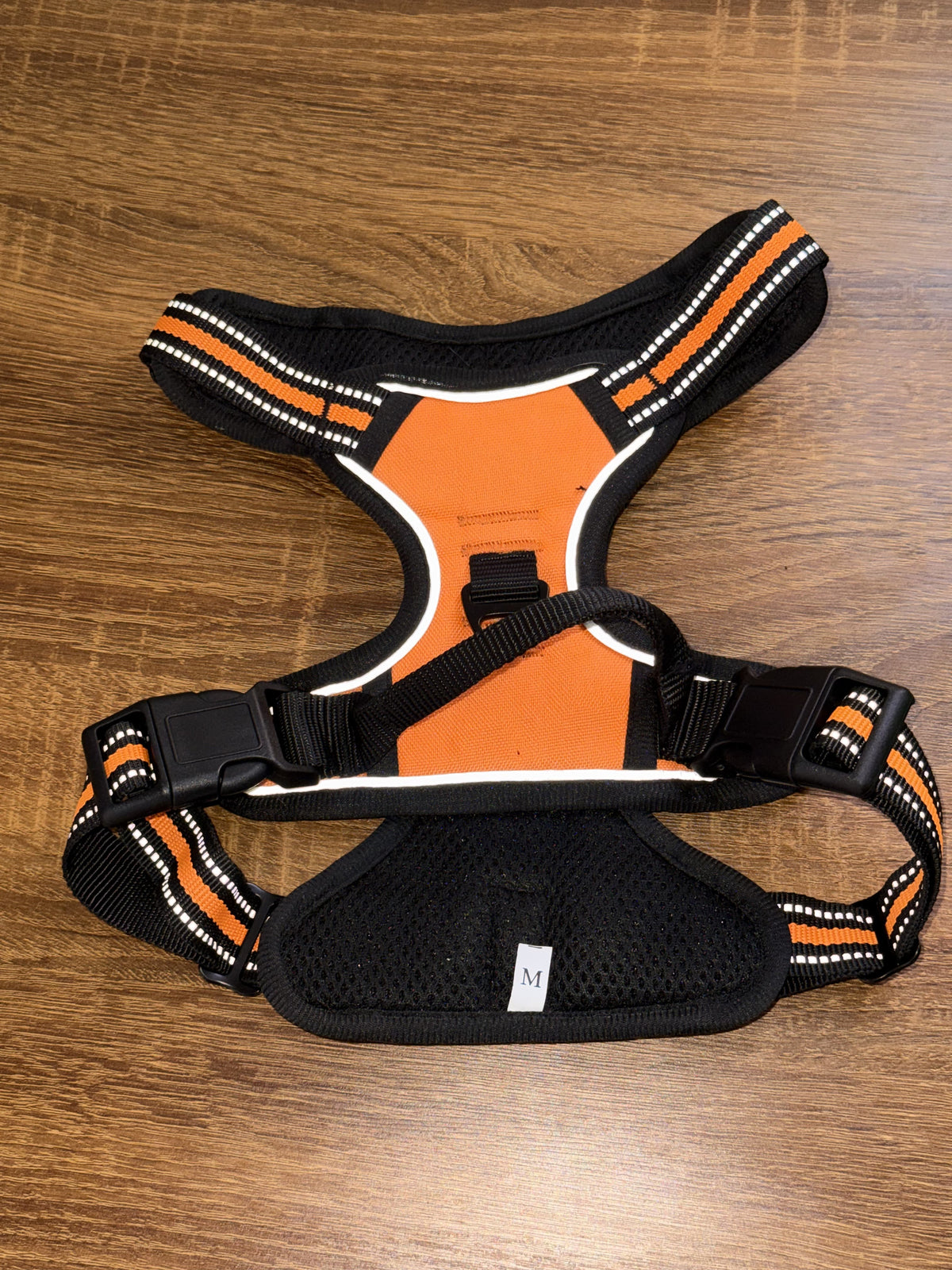 A black and orange dog harness with reflective striped straps, laid flat on a brown wooden surface.